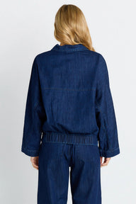 A woman viewed from the back wearing a dark blue denim jumpsuit with long sleeves and matching pants against a light gray studio background.