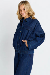 A woman wearing a dark blue denim jacket and matching denim pants, posing against a light gray studio background.