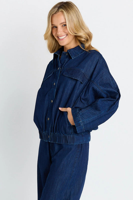 A woman wearing a dark blue denim jacket and matching denim pants, posing against a light gray studio background.