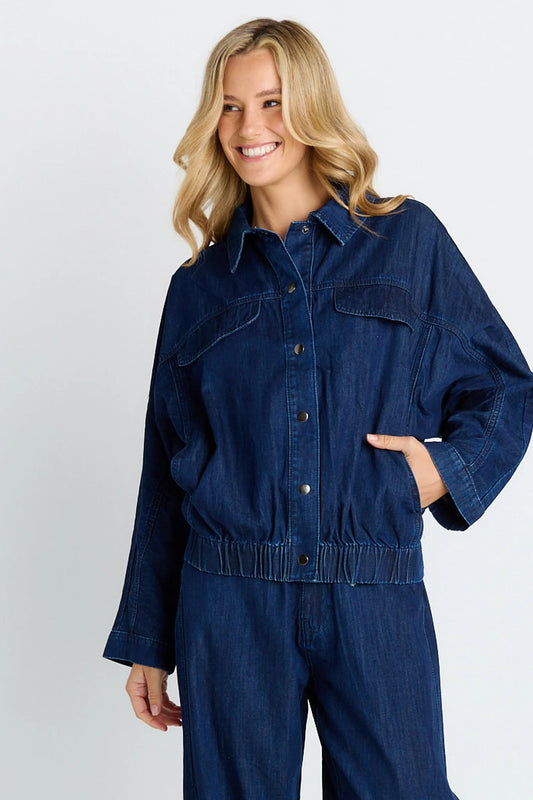 A woman wearing a dark blue denim jumpsuit with a button-up shirt-style top and matching pants, photographed against a light gray studio background.