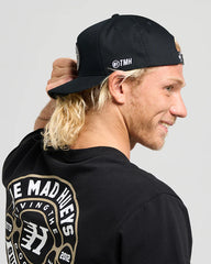 Person wearing a black cap with a logo and a black hoodie with gold text on the back, against a gray background.