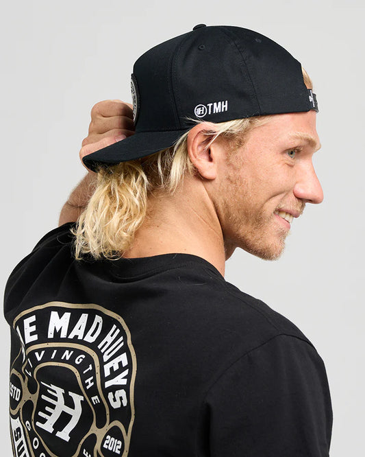 Person wearing a black cap with a logo and a black hoodie with gold text on the back, against a gray background.