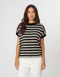 A woman modeling a short-sleeve knit top with horizontal black and beige stripes, paired with white pants, against a plain light gray studio background.