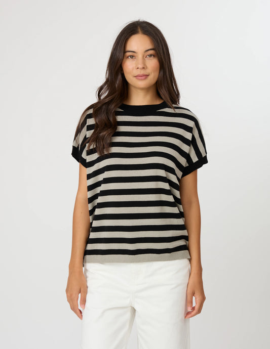 A woman modeling a short-sleeve knit top with horizontal black and beige stripes, paired with white pants, against a plain light gray studio background.
