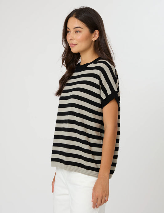A woman wearing a short-sleeve knit top with horizontal black and beige stripes, paired with white pants, photographed against a plain light gray studio background.
