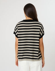 A woman viewed from behind wearing a short-sleeve knit top with horizontal black and beige stripes, paired with white pants, against a plain light gray studio background.