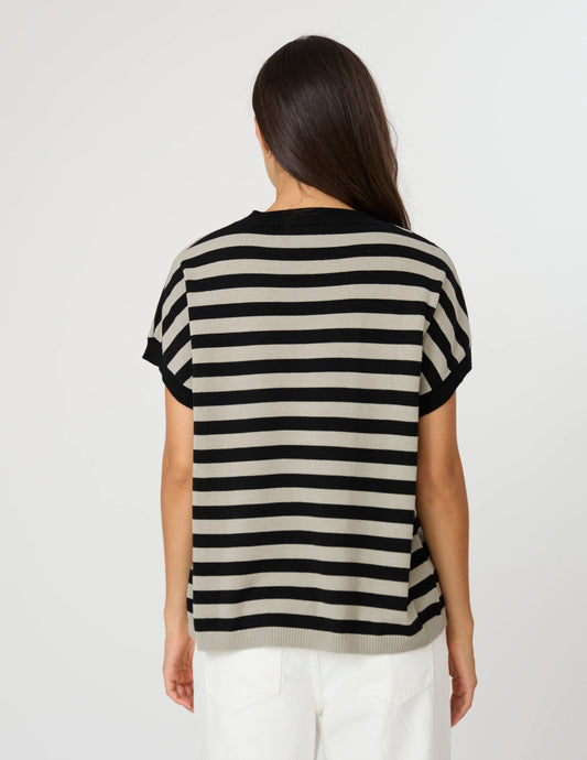 A woman viewed from behind wearing a short-sleeve knit top with horizontal black and beige stripes, paired with white pants, against a plain light gray studio background.