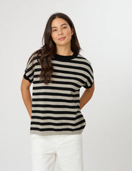 A woman modeling a short-sleeve knit top with horizontal black and beige stripes, paired with white pants, against a plain light gray studio background.