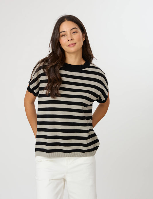 A woman modeling a short-sleeve knit top with horizontal black and beige stripes, paired with white pants, against a plain light gray studio background.