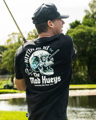 A man wearing a black t-shirt with a large graphic on the back, featuring a skull and the text 'HITTIN FK ALL THE MAD HUEYS'. He is outdoors near a pond with trees in the background.