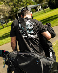 A man seen from behind wearing a black t-shirt with a large white and blue graphic on the back, carrying a black golf bag on his shoulder in an outdoor park setting.