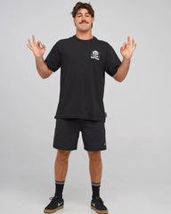 A man modeling a black short-sleeve t-shirt and matching black athletic shorts against a light gray studio background. The t-shirt features a small white graphic on the left chest, and the shorts have a small white logo near the hem.
