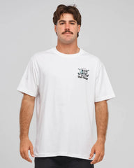 A man wearing a white short-sleeve crewneck t-shirt with a small black and white skull graphic and the text on the left chest, photographed against a solid light gray background.