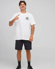 A man modeling a white crewneck t-shirt with a small black graphic on the left chest, paired with black athletic shorts, socks, and sneakers, against a light gray studio background.