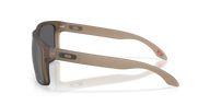 Oakley sunglasses with a translucent taupe-brown acetate frame and dark gray lenses, shown from a side profile on a white background with a soft reflection.