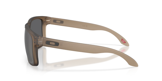 Oakley sunglasses with a translucent taupe-brown acetate frame and dark gray lenses, shown from a side profile on a white background with a soft reflection.