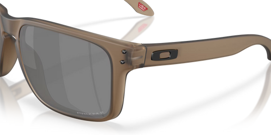 Oakley sunglasses with a translucent taupe-brown frame and dark gray lenses, shown on a clean white background with a soft reflection.