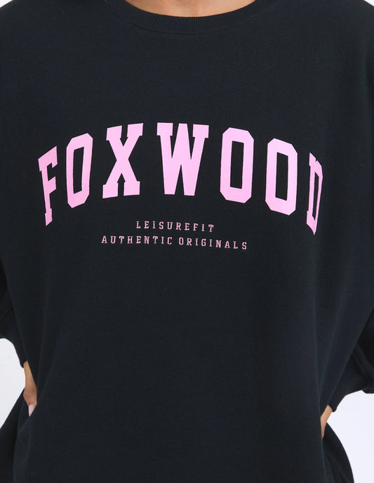 A close-up of a black crewneck sweatshirt featuring the word 'FOXWOOD' printed in large pink arched letters across the chest, with smaller pink text underneath reading 'LEISUREFIT AUTHENTIC ORIGINALS'.