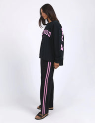 A woman modeling a black sweatshirt and matching track pants set with pink details, photographed against a plain white studio background.