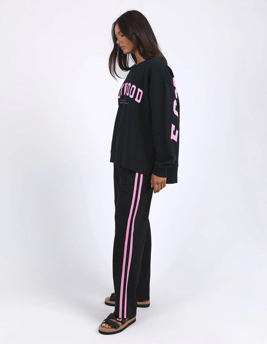 A woman modeling a black sweatshirt and matching track pants set with pink details, photographed against a plain white studio background.