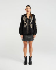Woman wearing a black embroidered blouse, black skirt, and black boots on a white background