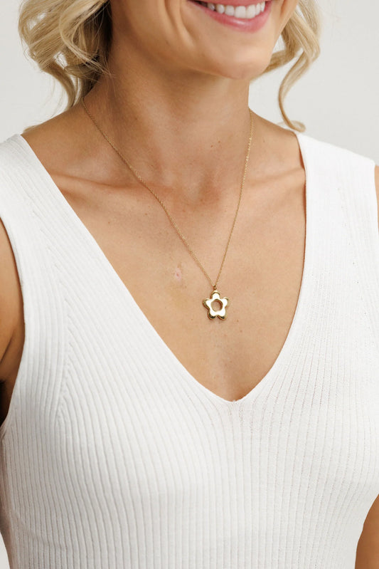 Woman wearing a white sleeveless top with a gold necklace featuring a flower pendant.