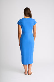 A woman viewed from the back wearing a bright blue short-sleeve midi dress, standing against a light gray studio background.