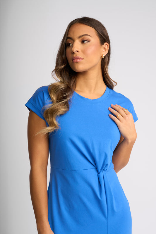 A woman wearing a bright blue short-sleeve dress with a gathered waist detail, posing against a light gray studio background.