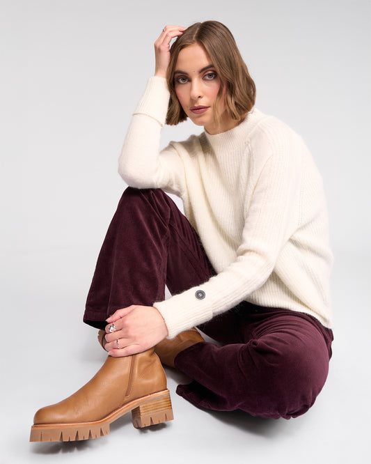 Woman wearing a white sweater and maroon pants with brown boots on a light gray background