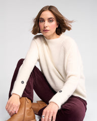 Woman wearing a white sweater and burgundy pants sitting on a chair with a plain background