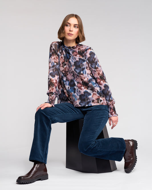 Woman sitting on a black stool wearing a floral blouse and blue jeans against a white background