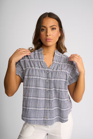 A woman modeling a short-sleeve button-up blouse with a blue, white, and gray gingham plaid pattern, paired with white pants, against a solid light gray background.