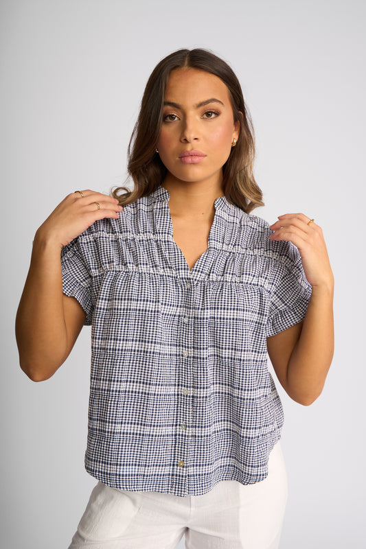 A woman modeling a short-sleeve button-up blouse with a blue, white, and gray gingham plaid pattern, paired with white pants, against a solid light gray background.