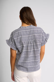 A woman viewed from the back wearing a short-sleeve plaid blouse with ruffled sleeves and white pants, against a light gray studio background.