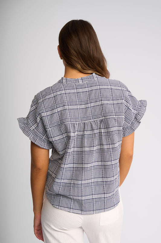 A woman viewed from the back wearing a short-sleeve plaid blouse with ruffled sleeves and white pants, against a light gray studio background.