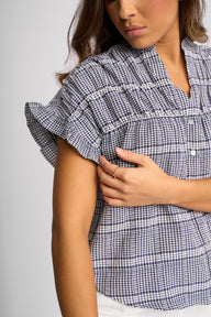 A close-up of a woman wearing a navy and white gingham short-sleeve blouse with ruffled sleeves and a smocked chest detail, paired with white bottoms.