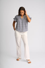 A woman modeling a short-sleeve plaid button-up shirt in blue and white, paired with off-white wide-leg pants and white sandals, against a light gray studio background.