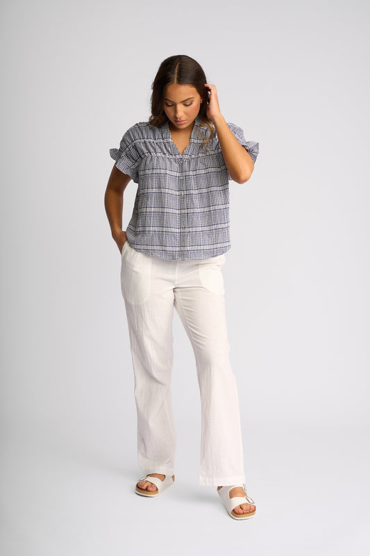 A woman modeling a short-sleeve plaid button-up shirt in blue and white, paired with off-white wide-leg pants and white sandals, against a light gray studio background.