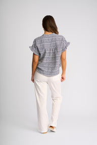 A woman viewed from behind wearing a gray and white plaid short-sleeve blouse with ruffled sleeves and white pants, standing against a light gray studio background.