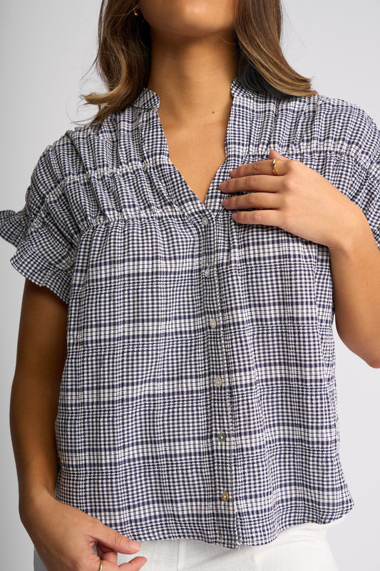 A woman wearing a short-sleeve button-front blouse with a navy and white gingham pattern and a smocked chest detail, paired with white bottoms against a light gray studio background.