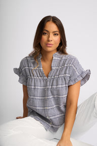 A woman wearing a short-sleeve button-up blouse with a blue and white gingham plaid pattern and ruffled sleeves, paired with white pants, seated against a light gray background.