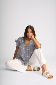 A woman sits on the floor wearing a blue and white plaid short-sleeve blouse, white wide-leg pants, and white slide sandals against a light gray studio background.