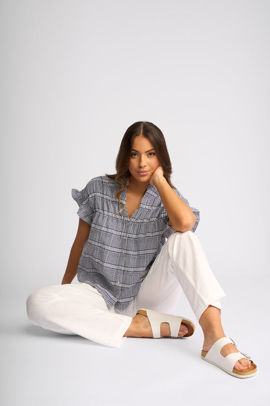 A woman sits on the floor wearing a blue and white plaid short-sleeve blouse, white wide-leg pants, and white slide sandals against a light gray studio background.
