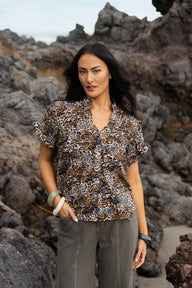 A woman wearing a short-sleeve button-up blouse with a brown, black, and white leopard print, paired with gray pants, standing on a rocky beach.