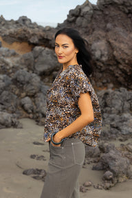 A woman stands on a rocky beach wearing a short-sleeve leopard-print blouse and gray pants, looking back over her shoulder.