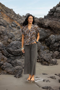 A woman stands barefoot on a sandy beach with dark volcanic rocks behind her, wearing a short-sleeve button-front blouse with a multicolor abstract print and wide-leg taupe pants.