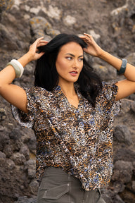 A woman posing outdoors in front of a rocky background, wearing a short-sleeve button-up blouse with a brown, black, and beige leopard print pattern, paired with gray pants.