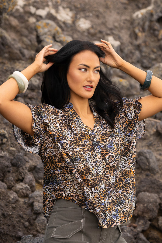 A woman posing outdoors in front of a rocky background, wearing a short-sleeve button-up blouse with a brown, black, and beige leopard print pattern, paired with gray pants.