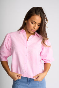 A woman wearing a light pink textured button-up blouse with puffed three-quarter sleeves and light blue jeans, standing against a light gray studio background.