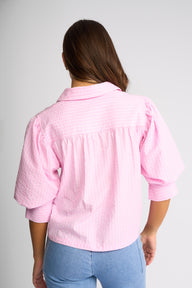 A woman viewed from the back wearing a light pink textured blouse with puff sleeves and light blue jeans, set against a plain light gray studio background.